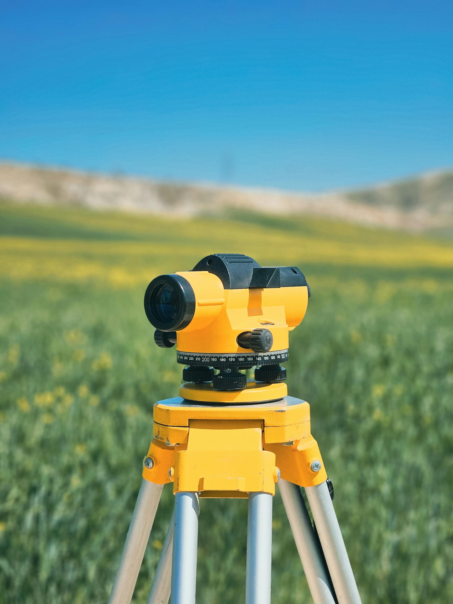 photo-54 A surveyor tripod with a yellow level in a green countryside field under a clear blue sky.